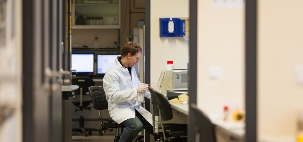 Foto: Ein Student der Biowissenschaften in einem Kittel gekleidet und mit blauen Füßlingen sitzt am Schreibtisch eines Labors