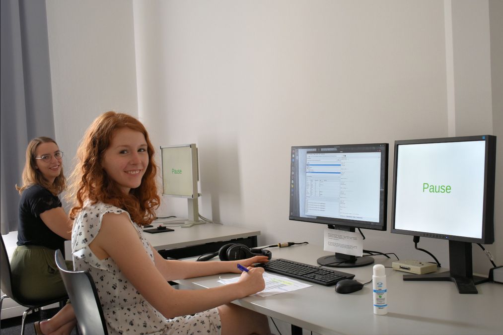 zur Vergrößerungsansicht des Bildes: Eine Versuchsleiterin und eine Versuchsperson sitzen in einem Computerlabor und lächeln in die Kamera. Auf dem Monitor ist das Wort „Pause“ zu sehen.