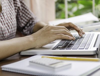 Eine Frau tippt auf einem Laptop. Neben ihr liegt ein Notizbuch und ein Stift.