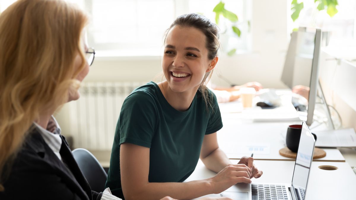 zur Vergrößerungsansicht des Bildes: Bild zeigt zwei Frauen, die vor einem Laptop sitzen und sich gegenseitig anlächeln