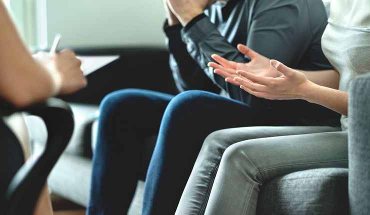 Ein Mann und eine Frau sitzen nebeneinander und gestikulierend auf einem Sofa. Ihnen gegenüber sitzt eine dritte Person. Diese macht Notizen auf einem Klemmbrett.