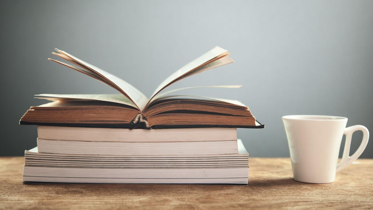 zur Vergrößerungsansicht des Bildes: Ein offenes Buch liegt auf mehreren geschlossenen Büchern. Daneben steht eine weiße Tasse.