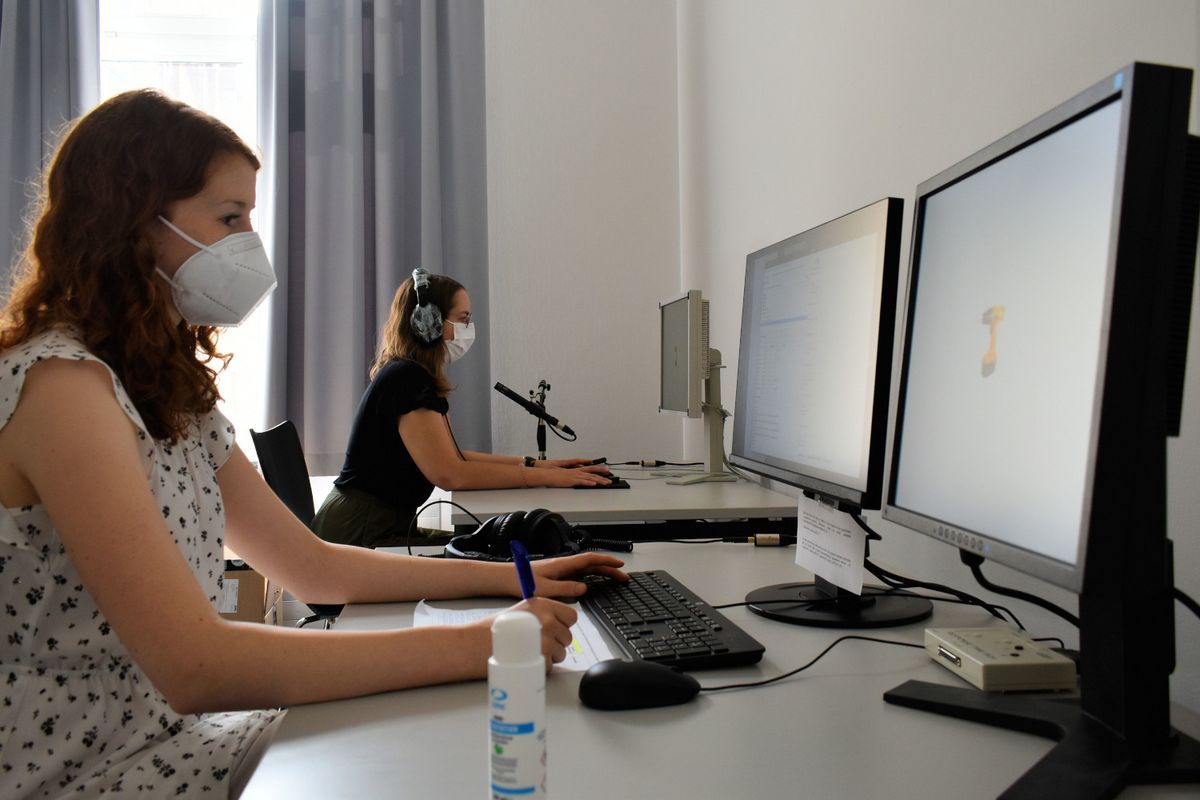 Eine Versuchsleiterin und eine Versuchsperson sitzen in einem Computerlabor und führen ein psychologisches Experiment zur Sprachverarbeitung durch. Beide tragen einen Mund-Nasenschutz.