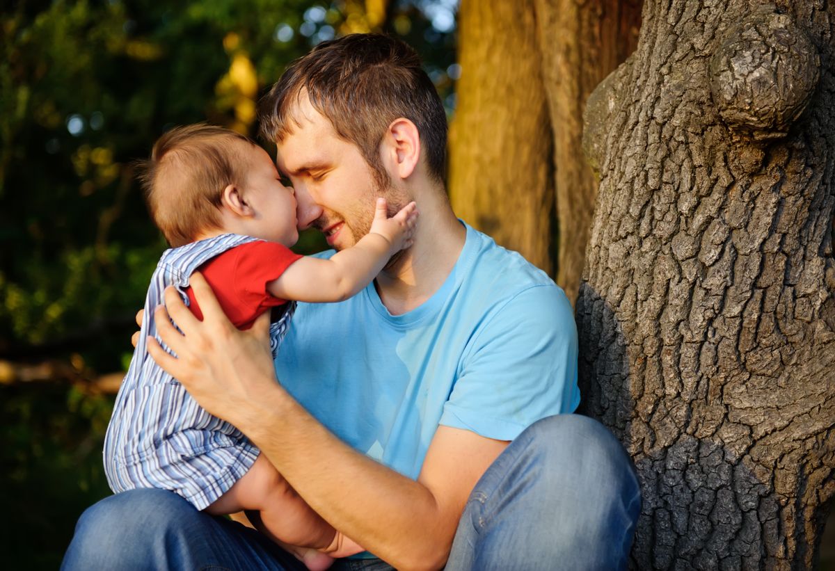 Ein Vater sitzt vor einem Baumstamm und hält zärtlich ein Baby im Arm.