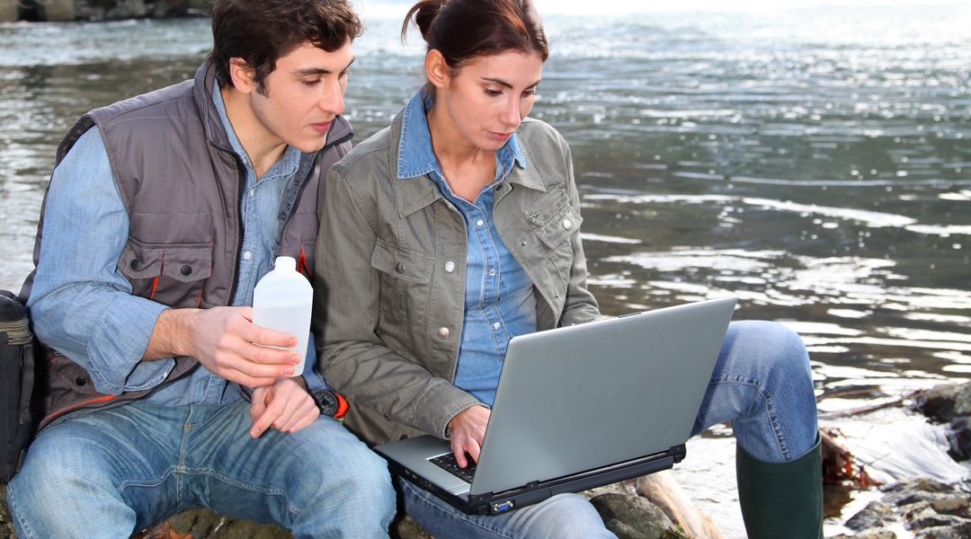 Ein junger Mann und eine junge Frau erheben am Flussufuer Wasserproben und erfassen die Ergebnisse in einem Laptop