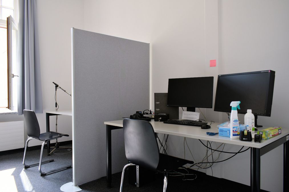 zur Vergrößerungsansicht des Bildes: Zu sehen ist ein Computerlabor mit zwei PC-Arbeitsplätzen (jeweils mit Tisch, Stuhl, Monitor, Tastatur, Maus, Kopfhörer und Mikrofon), die mit einer Stellwand voneinander getrennt sind.