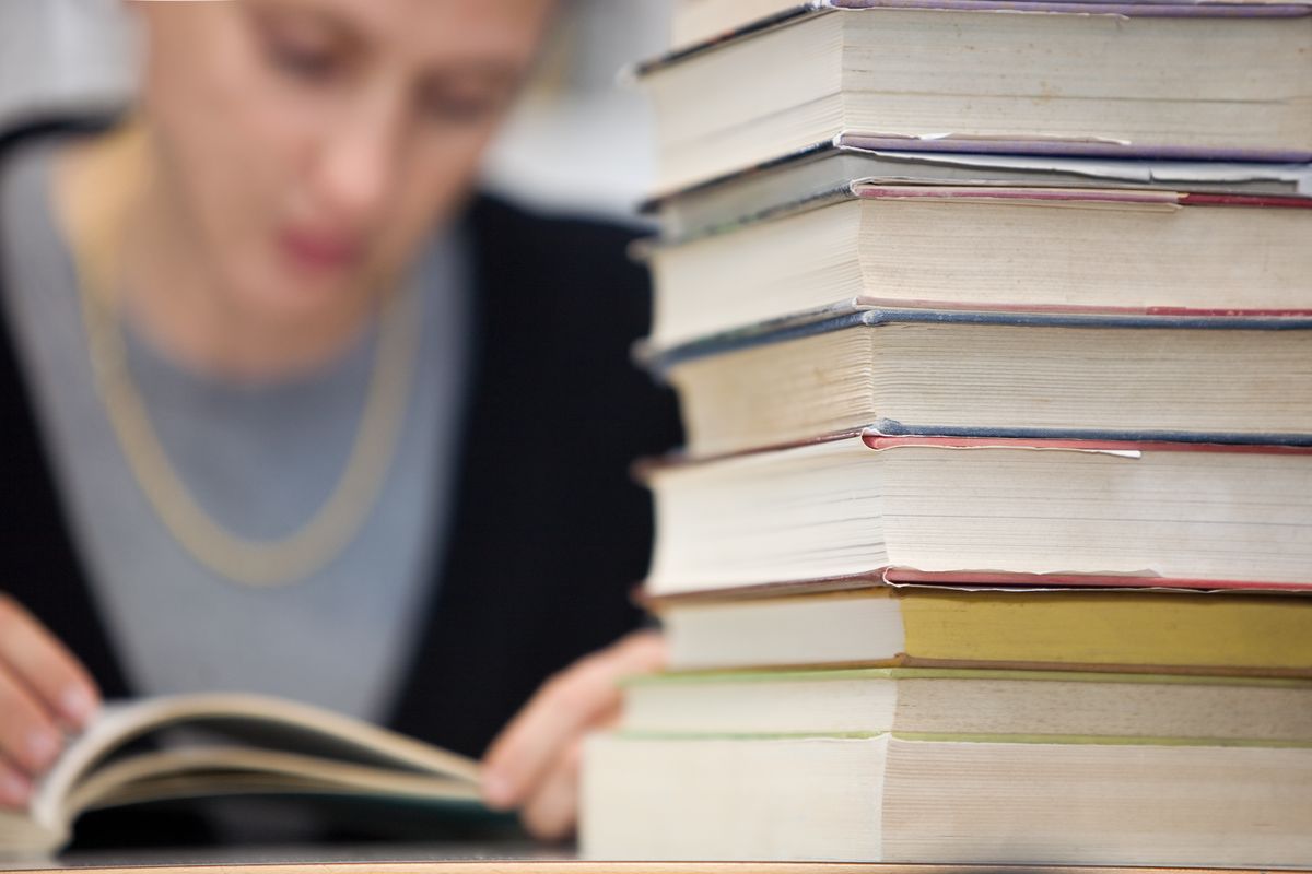 zur Vergrößerungsansicht des Bildes: Eine Studentin sitz an einem Tisch und liest in einem Buch, vor ihr liegt ein weiterer Stapel an Büchern.