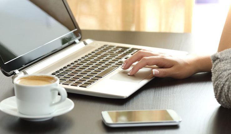 Laptop, Kaffee und Smartphone auf einem Tisch. Im Bildausschnitt zu sehen: Hand und Unterarm einer Frau sitzend vor dem Laptop.