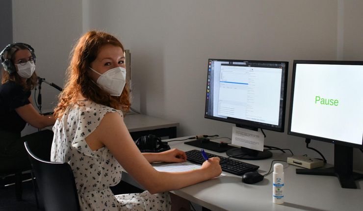 Eine Versuchsleiterin und eine Versuchsperson sitzen in einem Computerlabor und schauen in die Kamera. Beide tragen einen Mund-Nasenschutz. Auf dem Monitor ist das Wort „Pause“ zu sehen.